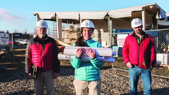 Three people, 2 men and a woman, standing outside of a building under construction. All are wearing hardhats, woman in the middle is holding rolled up building plans.